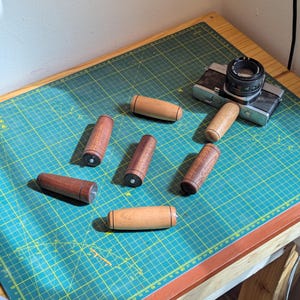 May include: Six wooden handles with a dark brown stain and a light brown wood grain. The handles are arranged on a green cutting mat with a silver camera in the background.