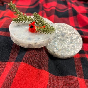 May include: Two round, decorative soap bars. One bar features a small red cardinal figurine and a sprig of green pine needles. The other bar is filled with silver and iridescent beads. The soaps are on a red and black plaid background.