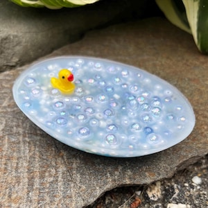 May include: An oval-shaped soap dish with a light blue, bubbly design. A small yellow rubber duck with a red beak floats in the center. The dish is set on a gray stone surface.