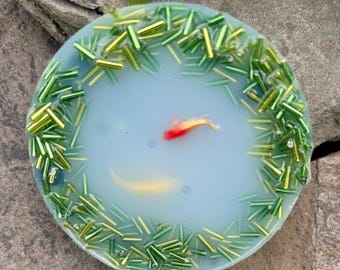 May include: A round, light blue resin pendant with a koi pond design. The pendant features a red and yellow koi fish, surrounded by green and gold beads, resembling aquatic plants. The pendant is set against a natural, textured background.