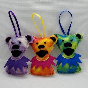 May include: Three tie-dye felt ornaments shaped like bears with different colored collars. The bears are purple, orange, and green.