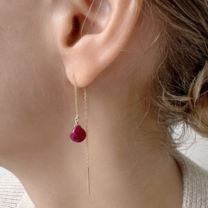 May include: Gold chain earrings with a dangling red gemstone. The earrings are shown on a person's ear.