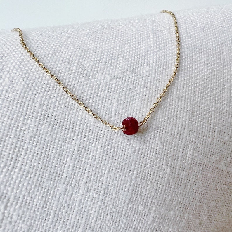 May include: A fine gold chain necklace with a single, faceted, deep red gemstone pendant. The necklace is displayed on a white, textured surface, contrasting the gold and the gemstone.