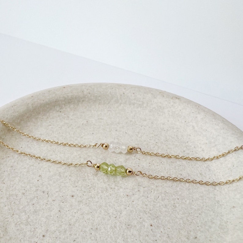 Op de afbeelding: Twee delicate gouden kettingen met edelsteen accenten. De bovenste ketting heeft witte kralen, terwijl de onderste ketting groene kralen heeft.