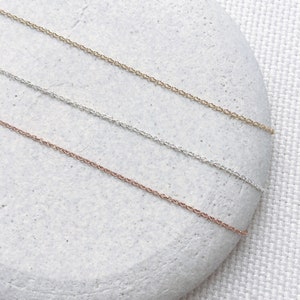 May include: Three delicate chain necklaces in gold, silver, and rose gold, displayed on a light grey stone background.