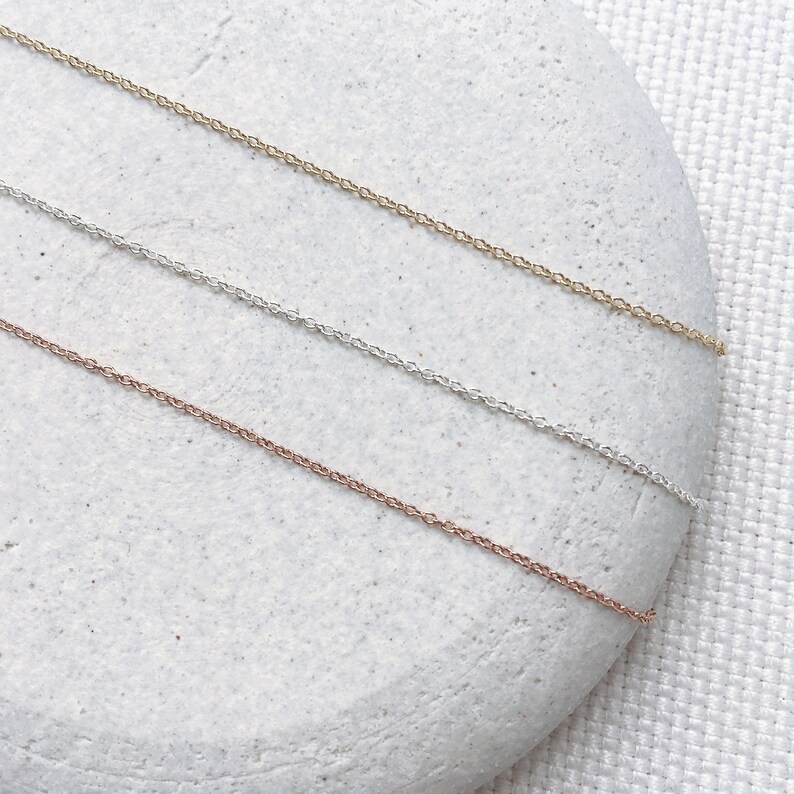 May include: Three delicate chain necklaces in gold, silver, and rose gold, displayed on a light grey stone background.