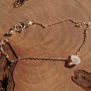 May include: A delicate gold chain necklace featuring a small white pearl and a larger, clear, faceted gemstone pendant.