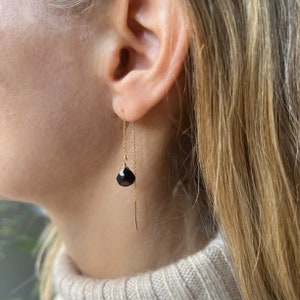 May include: A close-up of a gold chain earring with a black teardrop-shaped stone. The earring is worn in the ear of a person with blonde hair and a beige sweater. The earring hangs down with a thin gold bar at the end.