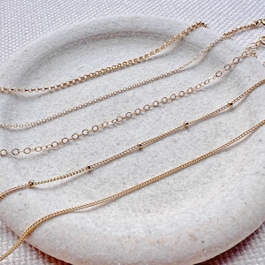 May include: Five gold chains of different styles and lengths, displayed on a white surface. The chains are all delicate and feature a variety of links and beads.
