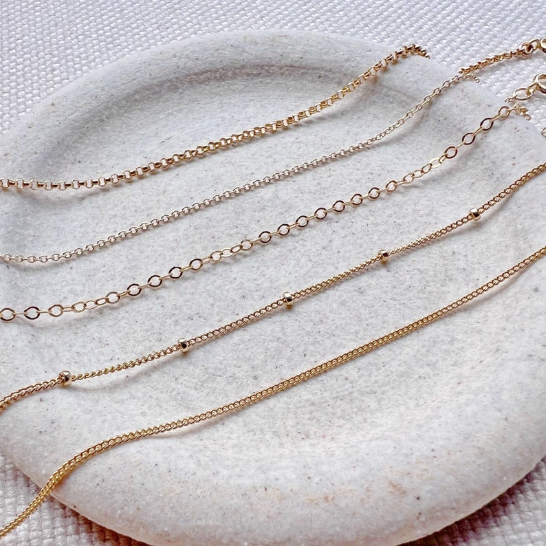 May include: Five gold chains of different styles and lengths, displayed on a white surface. The chains are all delicate and feature a variety of links and beads.