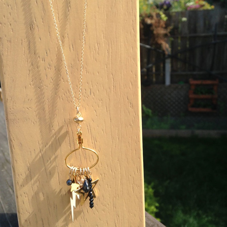 May include: A gold chain necklace with a gold charm pendant. The charm is a circle with a smaller circle inside. Hanging from the smaller circle are several gold stars, black beads, and a gold lightning bolt charm.