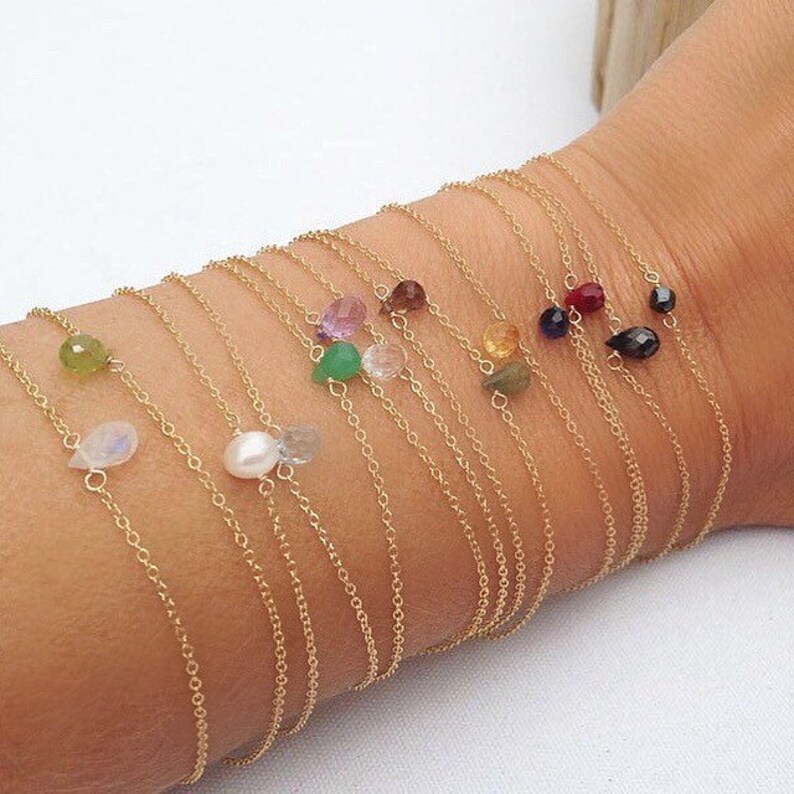 May include: A collection of delicate gold chain bracelets with small gemstone accents. Each bracelet features a different colored gemstone, including green, blue, red, purple, and white.