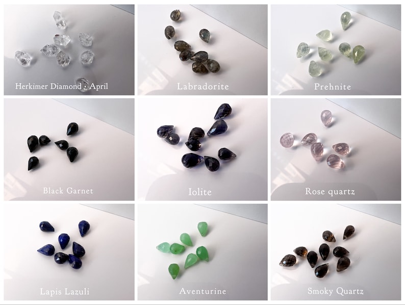 May include: Assortment of gemstone teardrop beads in various colours, including clear, black, blue, green, brown, and pink. The beads are arranged in rows on a white surface. The names of each gemstone are listed below each row.