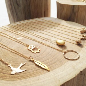 May include: A display of gold necklaces with various charms, including a bird, feather, owl, circle, and a textured stone. The necklaces are displayed on a wooden surface.