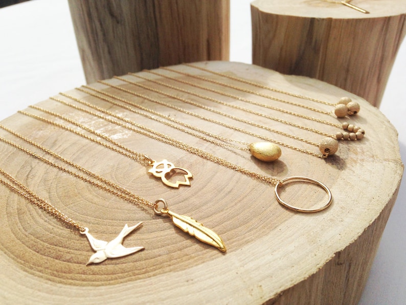 May include: A display of gold necklaces with various charms, including a bird, feather, owl, circle, and a textured stone. The necklaces are displayed on a wooden surface.