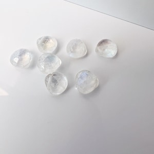 May include: Seven iridescent white moonstone cabochons, each with a unique, faceted shape, are arranged on a white surface.