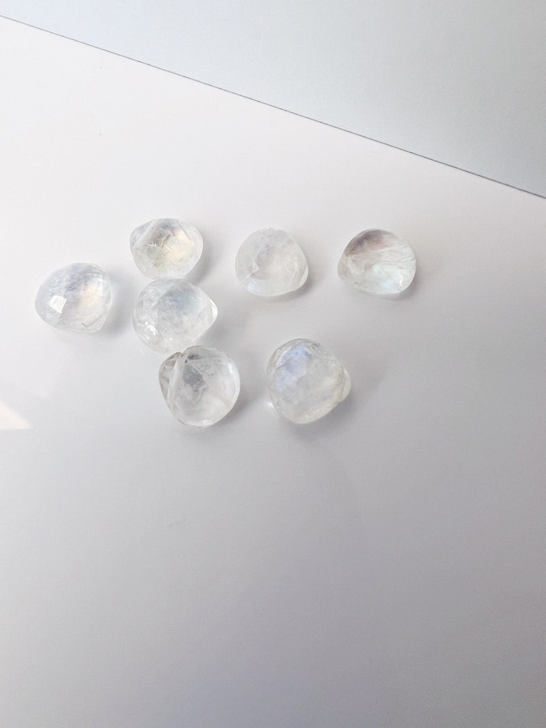 May include: Seven iridescent white moonstone cabochons, each with a unique, faceted shape, are arranged on a white surface.