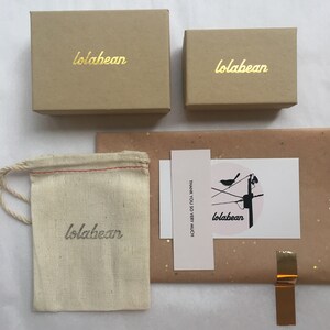 May include: A brown box with the text "lolabean" in gold lettering. The box is sitting on a brown surface with a white card that says "THANK YOU SO VERY MUCH" and a pink circle with the text "lolabean" and a black bird on a wire.
