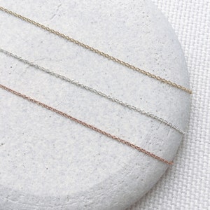 May include: Three delicate chain necklaces in gold, silver, and rose gold. The chains are displayed on a white stone background.