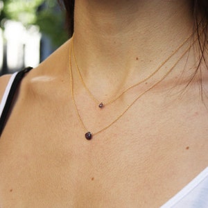 Puede incluir: Una mujer con un top blanco sin mangas y dos collares de oro con peque&ntilde;as piedras preciosas moradas.