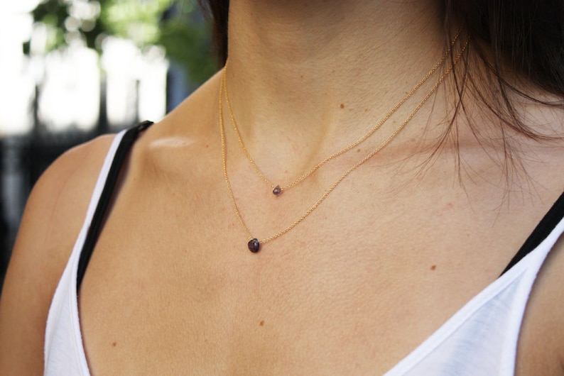 Puede incluir: Una mujer con un top blanco sin mangas y dos collares de oro con peque&ntilde;as piedras preciosas moradas.