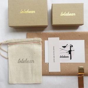 May include: A brown cardboard box with the text "lolabean" printed in gold lettering. The box is sitting on a white surface. There is a smaller brown cardboard box with the same text in gold lettering. There is a white cloth bag with the same text in black lettering. There is a white card with the text "THANK YOU SO VERY MUCH" and a black and white illustration of a bird on a wire with the text "lolabean" in black lettering.