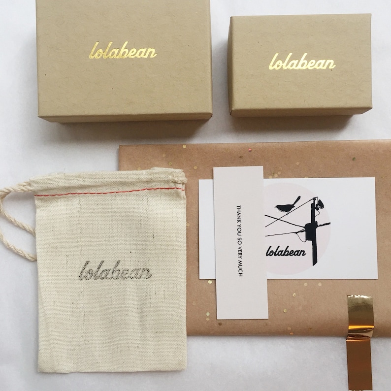 May include: A brown cardboard box with the text "lolabean" printed in gold lettering. The box is sitting on a white surface. There is a smaller brown cardboard box with the same text in gold lettering. There is a white cloth bag with the same text in black lettering. There is a white card with the text "THANK YOU SO VERY MUCH" and a black and white illustration of a bird on a wire with the text "lolabean" in black lettering.