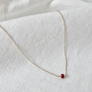 May include: A fine gold-coloured necklace with a small, cylindrical, deep red gemstone pendant. The chain is delicate and elegant. The necklace is displayed on a white, textured surface.