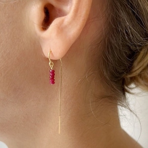 May include: A close-up of a gold earring with a chain and a cluster of small, red gemstones. The earring is dangling from the ear. The earring is a delicate piece of jewelry.