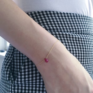 May include: A delicate gold chain bracelet with a single teardrop-shaped red gemstone pendant.