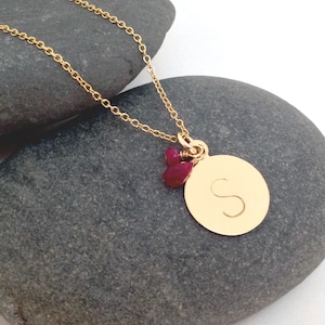 Op de afbeelding: Gouden ketting met een ronde hanger met de letter "S" en twee kleine rode edelstenen.