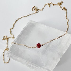 May include: A fine gold chain necklace with a single, faceted, deep red ruby gemstone pendant. The necklace is displayed on a white surface, highlighting its minimalist design and elegant simplicity.