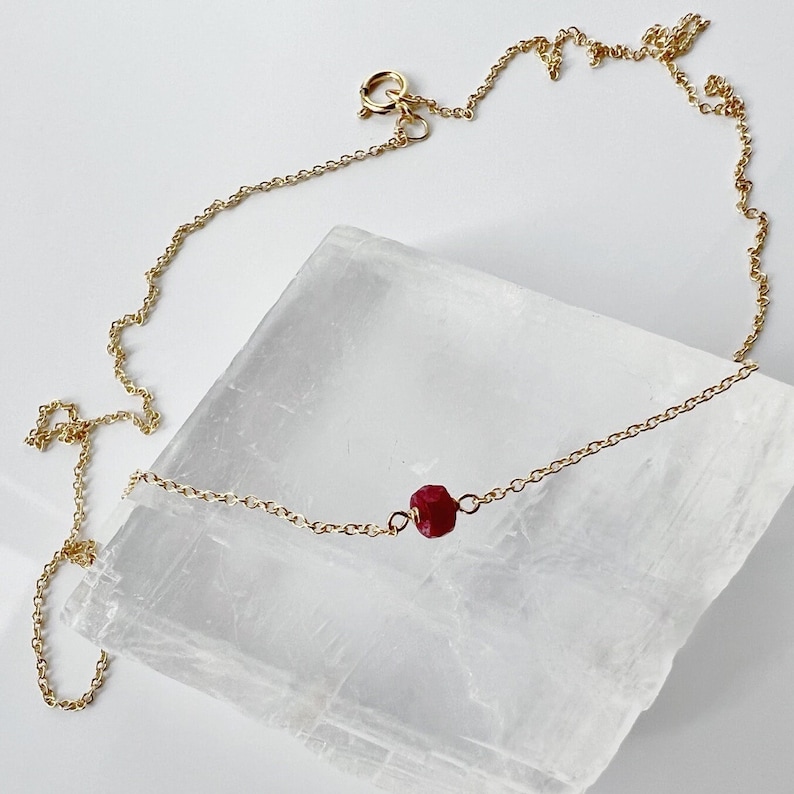 May include: A fine gold chain necklace with a single, faceted, deep red ruby gemstone pendant. The necklace is displayed on a white surface, highlighting its minimalist design and elegant simplicity.