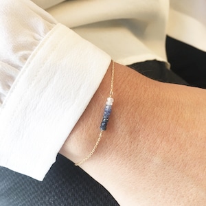K&ouml;nnte beinhalten: Ein zartes Armband aus einer goldenen Kette mit einer abgestuften Reihe von blauen und wei&szlig;en Edelsteinen. Das Armband wird am Handgelenk einer Person gezeigt.