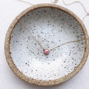 Pu&ograve; includere: Una delicata collana a catena dorata con un ciondolo di pietra preziosa rosa. La collana &egrave; esposta in una ciotola di ceramica bianca con macchie marroni.