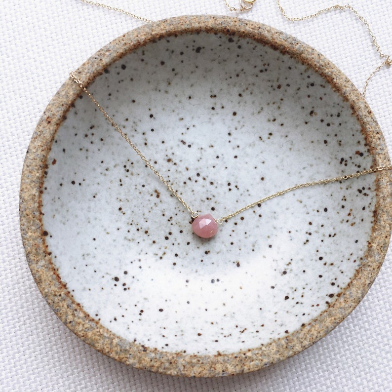 Pu&ograve; includere: Una delicata collana a catena dorata con un ciondolo di pietra preziosa rosa. La collana &egrave; esposta in una ciotola di ceramica bianca con macchie marroni.