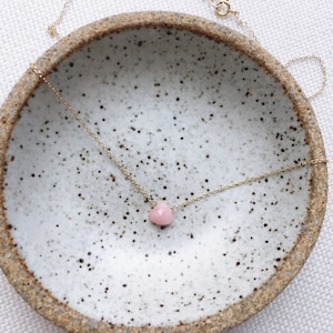 Pu&ograve; includere: Una delicata collana a catena d'oro con un ciondolo di pietra preziosa rosa. La collana &egrave; esposta su un piatto in ceramica macchiato.