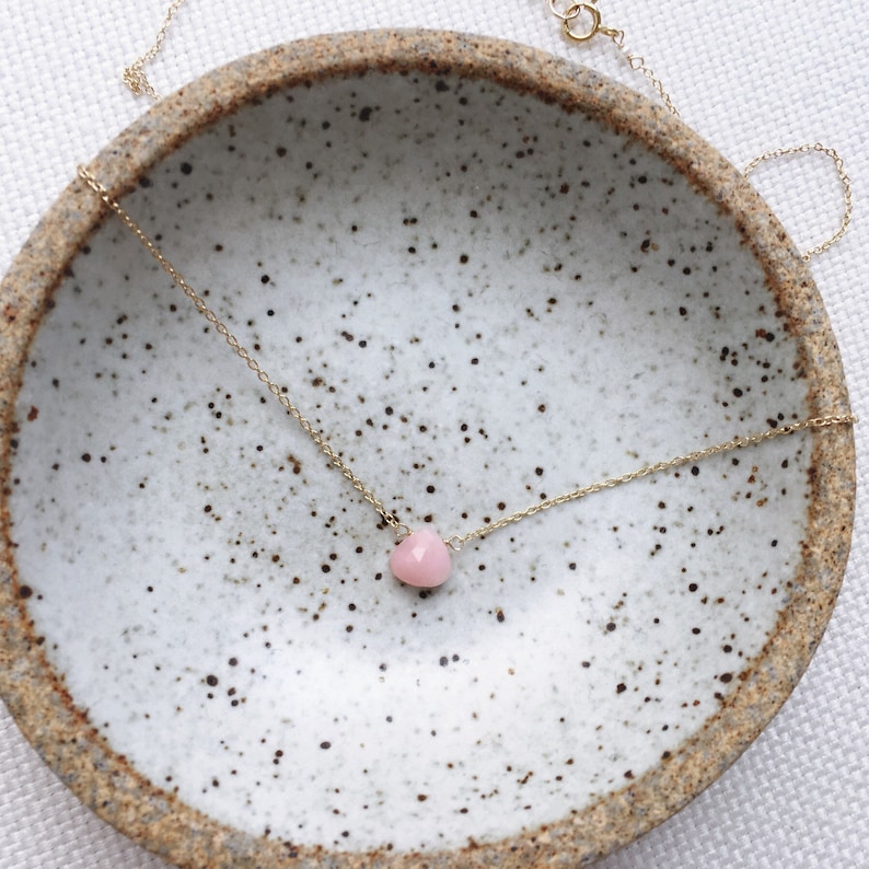 Pu&ograve; includere: Una delicata collana a catena d'oro con un ciondolo di pietra preziosa rosa. La collana &egrave; esposta su un piatto in ceramica macchiato.