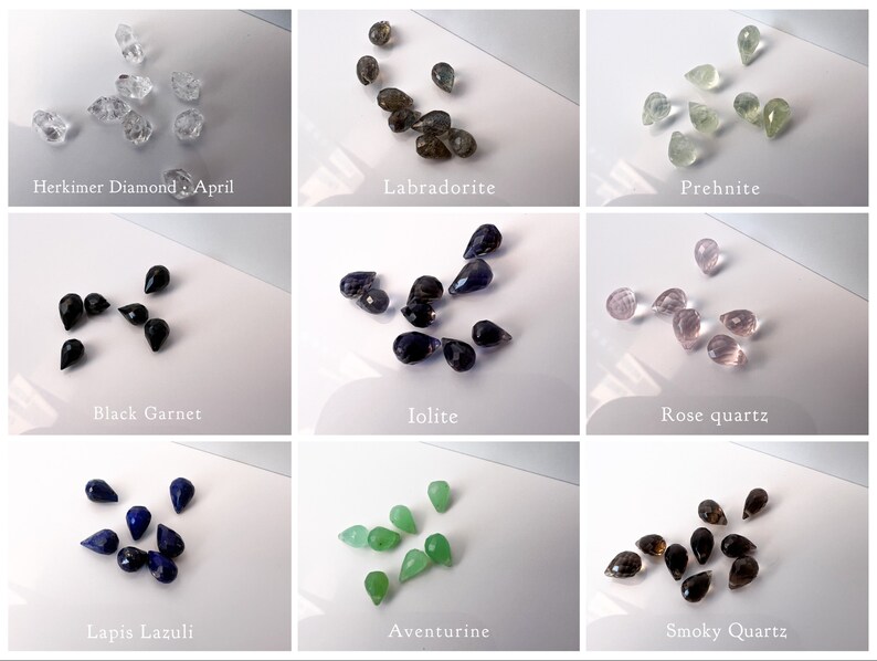 May include: Assortment of gemstone teardrop beads in various colors, including clear, black, blue, green, brown, and pink. The beads are arranged in rows on a white surface. The names of each gemstone are listed below each row.