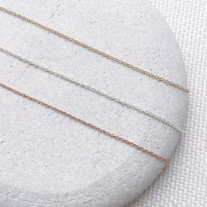 May include: Three delicate chain necklaces in gold, silver, and rose gold. The chains are laid out on a light gray stone background.