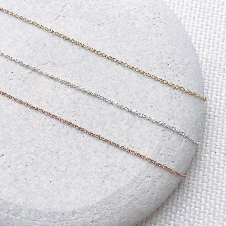 May include: Three delicate chain necklaces in gold, silver, and rose gold. The chains are laid out on a light gray stone background.