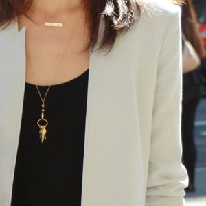 May include: A woman wearing a white blazer over a black top with a gold necklace featuring a charm with multiple keys.