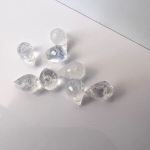 May include: A group of eight clear teardrop-shaped crystal beads. The beads are faceted and have a slight iridescent sheen.