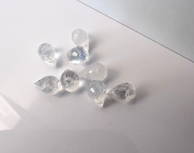 May include: A group of eight clear teardrop-shaped crystal beads. The beads are faceted and have a slight iridescent sheen.