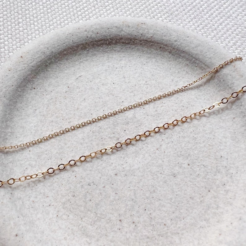 May include: Two gold chains on a white surface. The chains are different lengths and have different link styles.