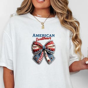 May include: White t-shirt with a patriotic bow design in red, white, and blue. The text "AMERICAN Sweetheart" is above the bow. The shirt has a relaxed, oversized fit, suitable for casual wear.