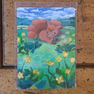 May include: A hand-painted Pokemon card featuring a brown and orange Vaporeon in a field of yellow flowers. The card has a blue sky with white clouds in the background.