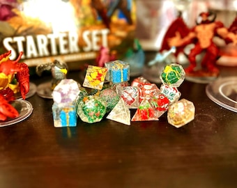 May include: A collection of colorful, decorative dice, some with embedded designs, are displayed on a dark surface. The dice are in various shapes, including cubes and polyhedrons. A box labeled "STARTER SET" is visible in the background, along with miniature figurines.