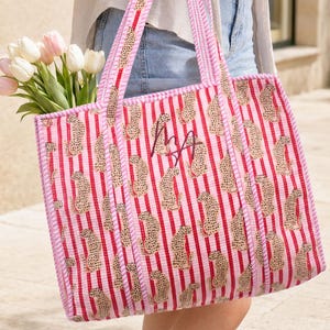 May include: A quilted tote bag featuring a pattern of vertical red stripes and small tan leopard figures. The bag has two long shoulder straps with a pink and white striped trim and a dark embroidered monogram in the center, filled with tulips.