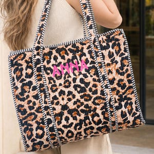 May include: A large rectangular tote bag featuring a tan and black leopard print pattern. The bag has black and white striped fabric handles and trim. The name ANNA is embroidered in bright pink thread on the front center of the bag.